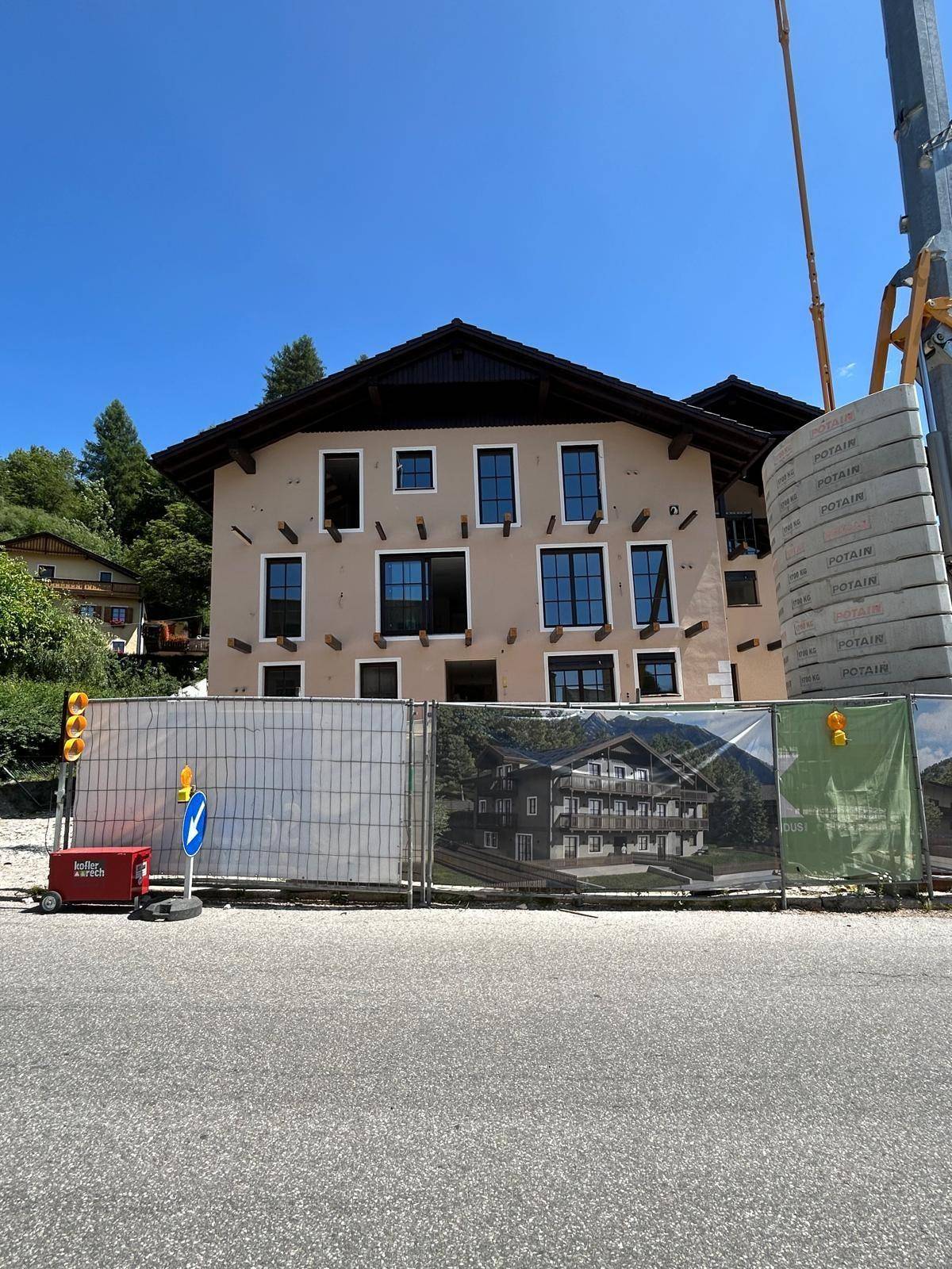 Vorderansicht des Neubaus von der Straße aus, mit Bauzaun und Visualisierung des fertigen Gebäudes.