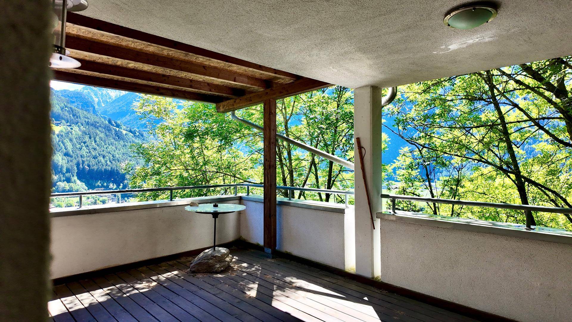 Überdachter Balkon mit Holzboden und einem kleinen Tisch, umgeben von grünen Bäumen und Bergblick.