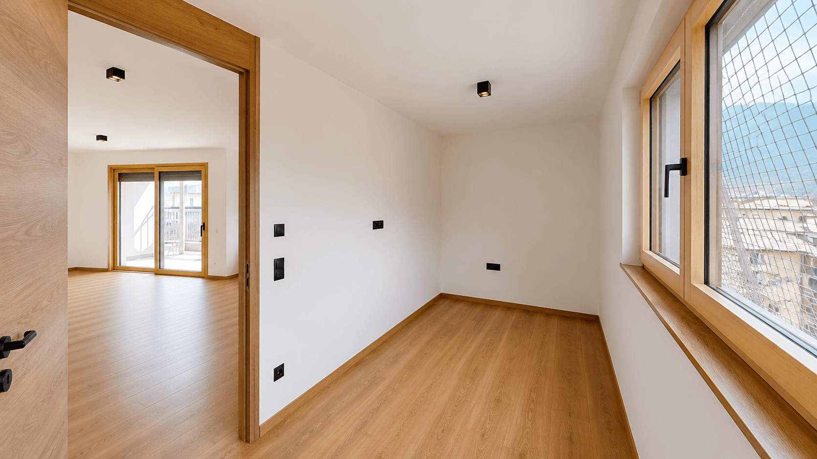 Helles Zimmer mit Holzboden und Fenster, das in einen weiteren Raum mit Balkonzugang führt.