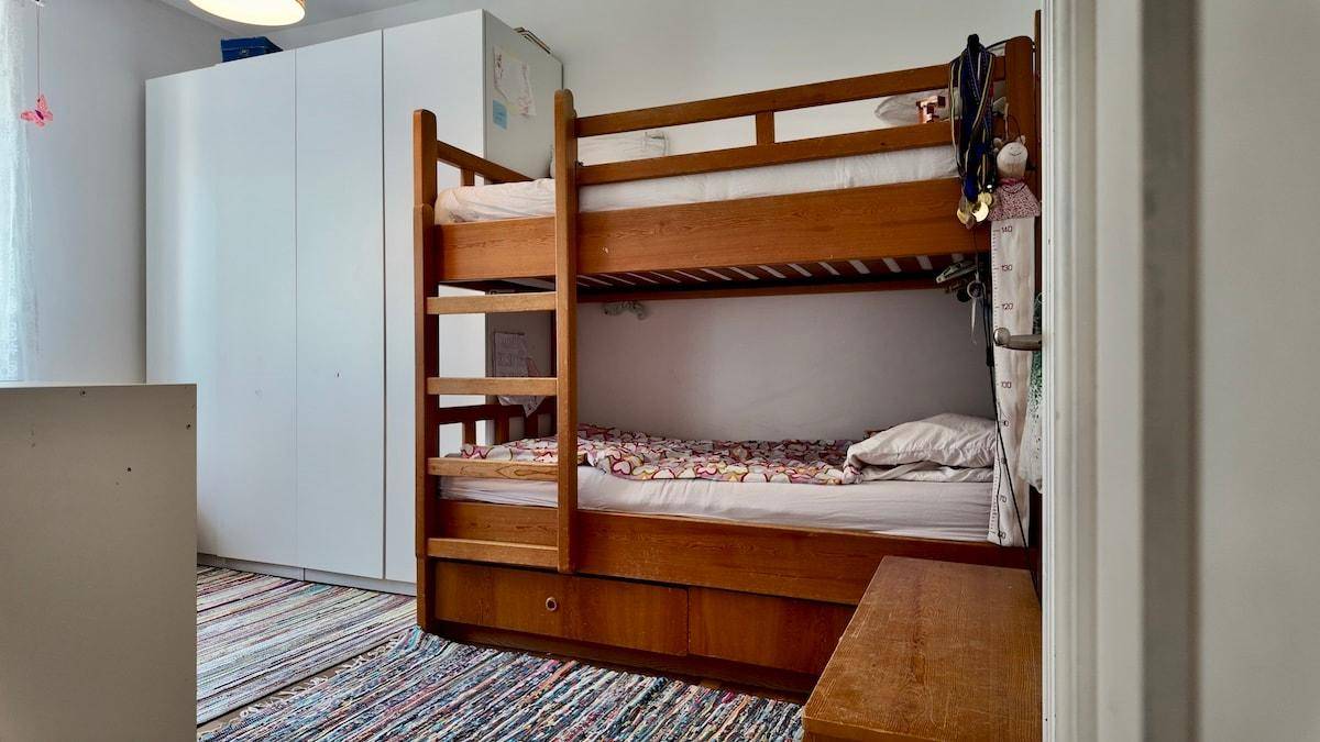 Kinderzimmer mit Etagenbett aus Holz und weißem Kleiderschrank, auf einem bunten Teppich.