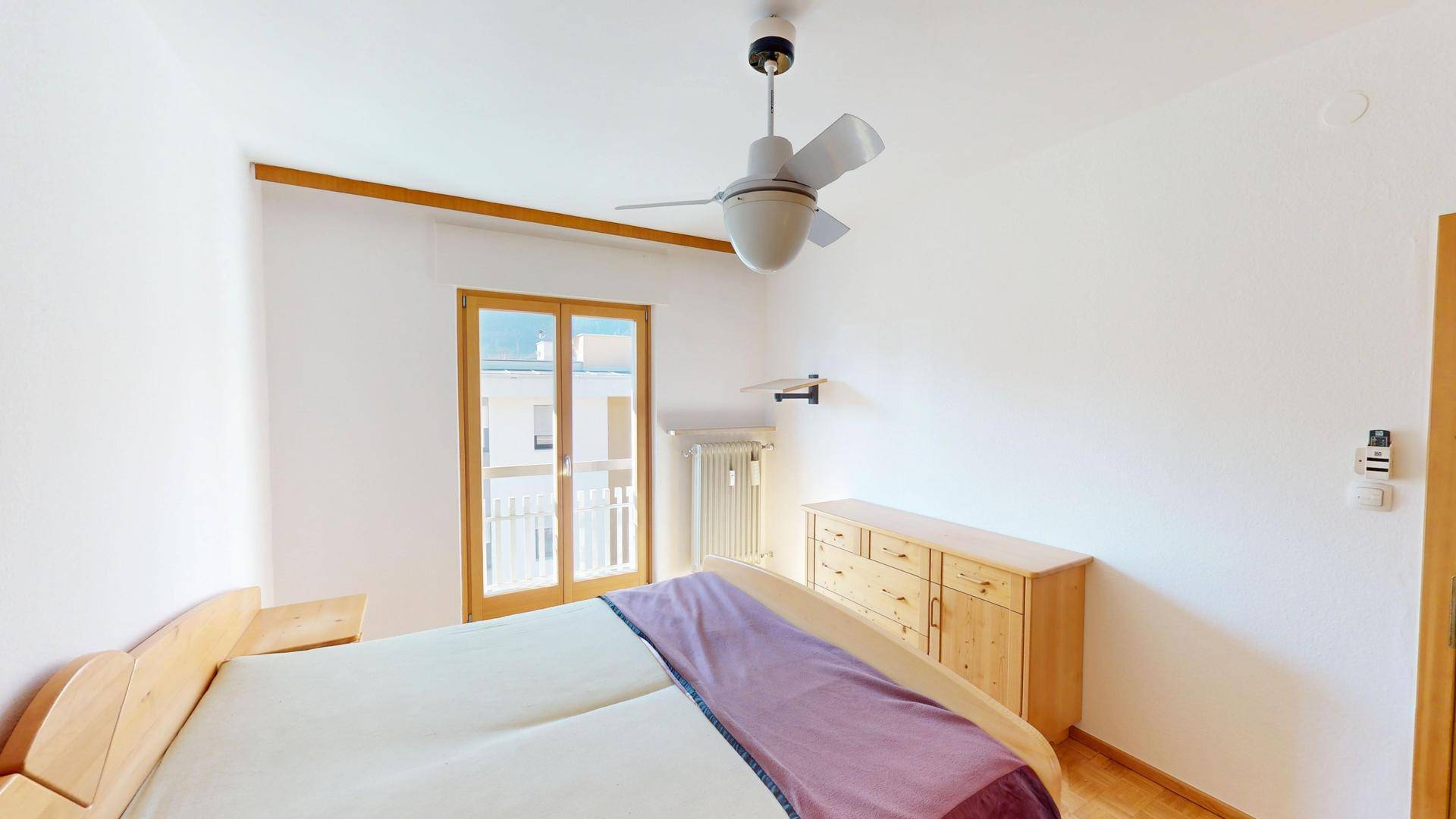 Schlafzimmer mit Holzbett, Kommode und Zugang zu einem kleinen Balkon.