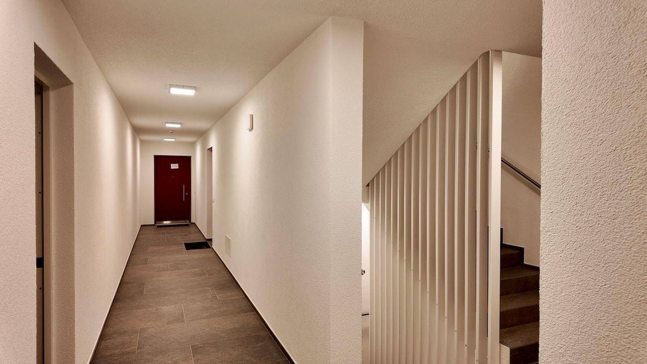 Langer Flur mit weißen Wänden, Fliesenboden und einer modernen Treppe mit Geländer.