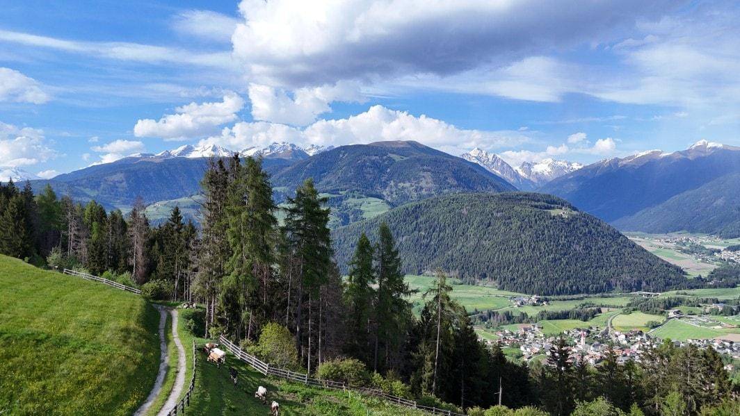 Panoramablick über ein grünes Tal mit einem Dorf, umgeben von bewaldeten Hügeln und schneebedeckten Bergen.