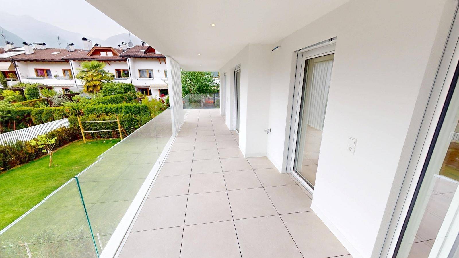 Geräumiger Balkon mit Fliesenboden, Glasgeländer und Blick auf die grüne Umgebung und Berge.