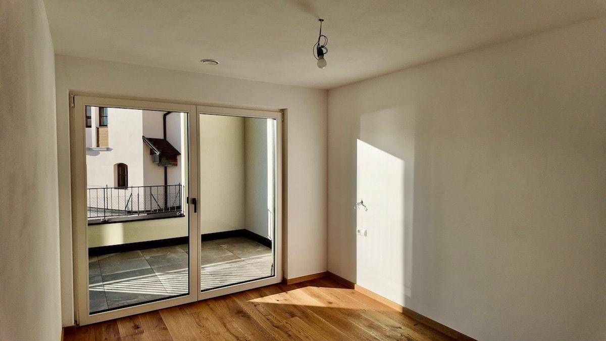Geräumiges Zimmer mit Holzboden und großer Schiebetür, die auf einen Balkon führt.