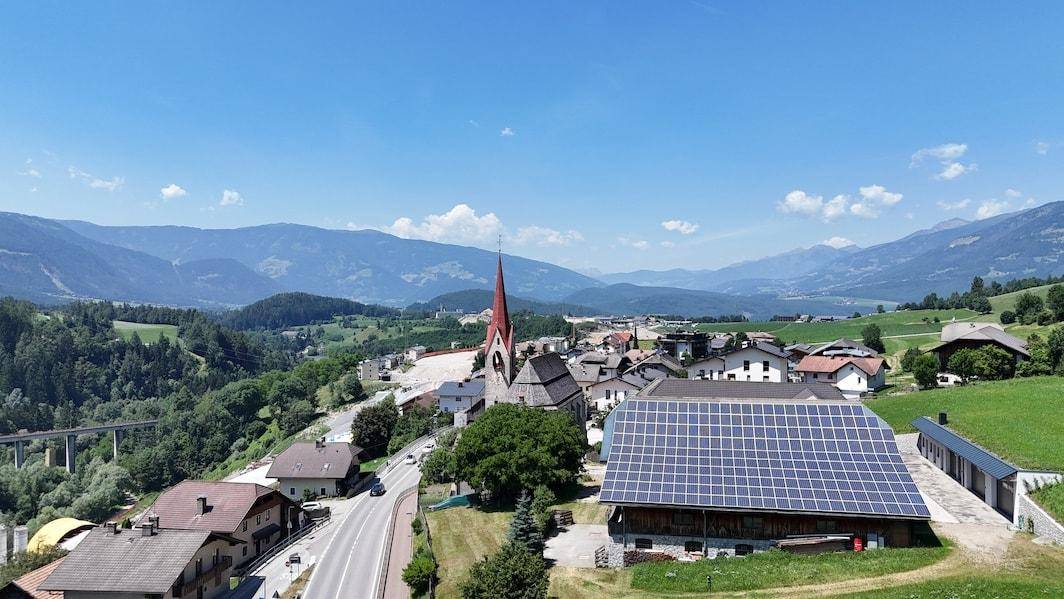 Panoramablick auf ein Bergdorf mit Kirche, großen Solaranlagen auf einem Dach und einer Straße im Tal.