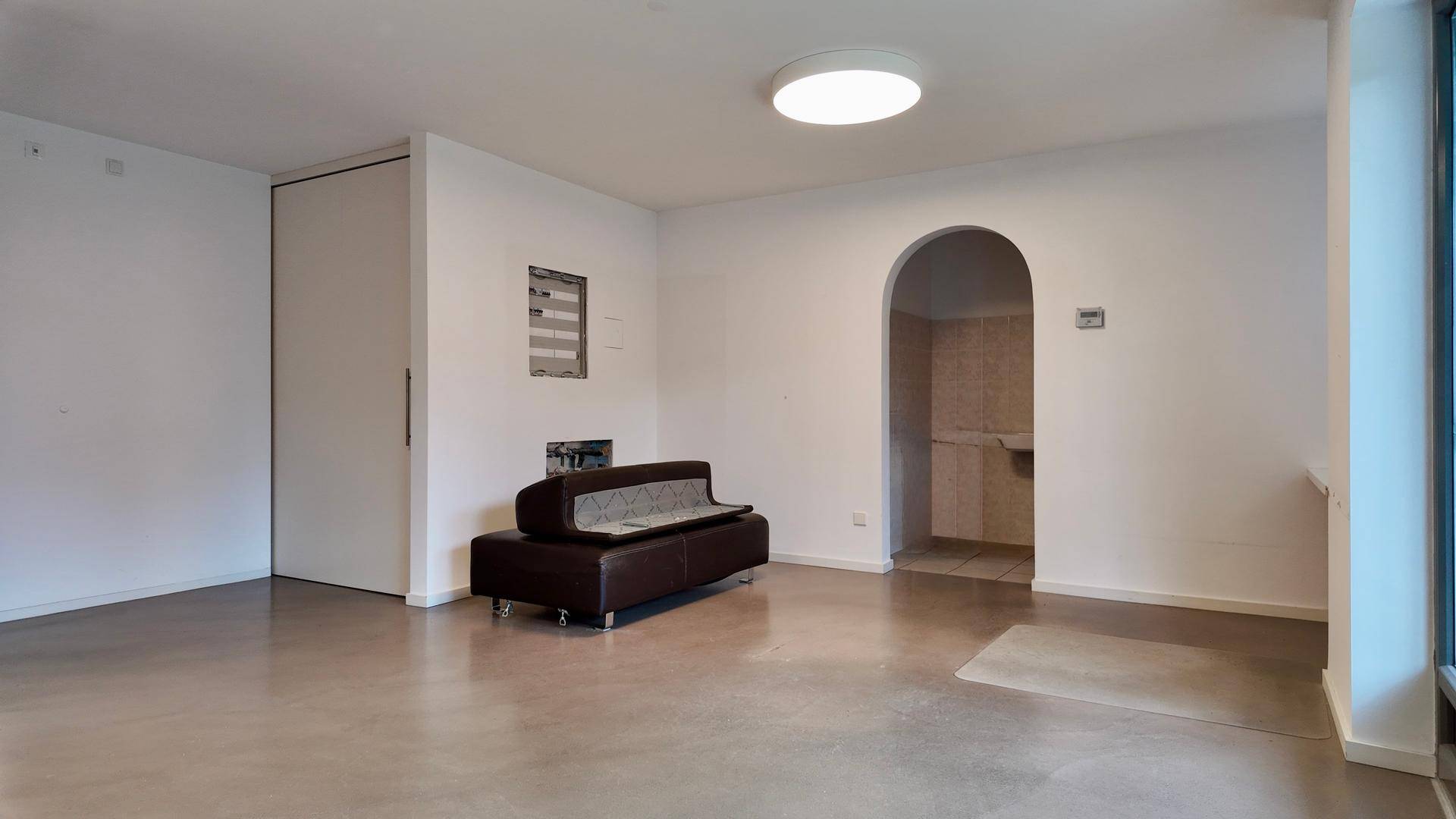 Geräumiger Wohnbereich mit einem dunklen Sofa, hellen Wänden und einem Bogendurchgang zu einem Nebenraum.