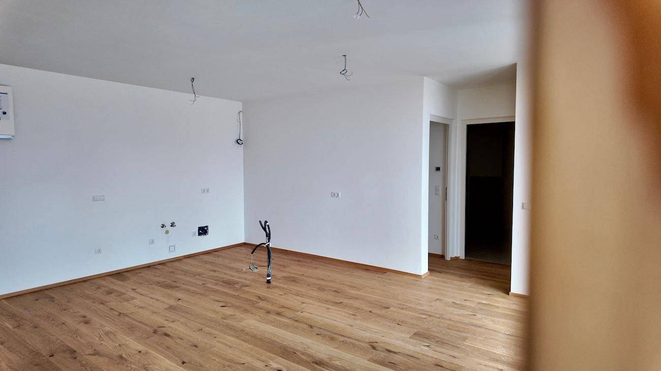 Leerer Raum mit neuem Holzboden, weißen Wänden und sichtbaren Elektroinstallationen.
