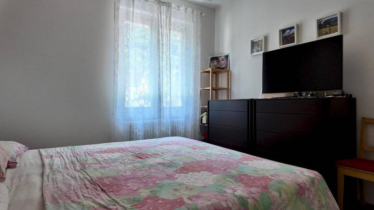 Schlafzimmer mit Doppelbett, Kommoden und Fernseher, beleuchtet durch ein großes Fenster.