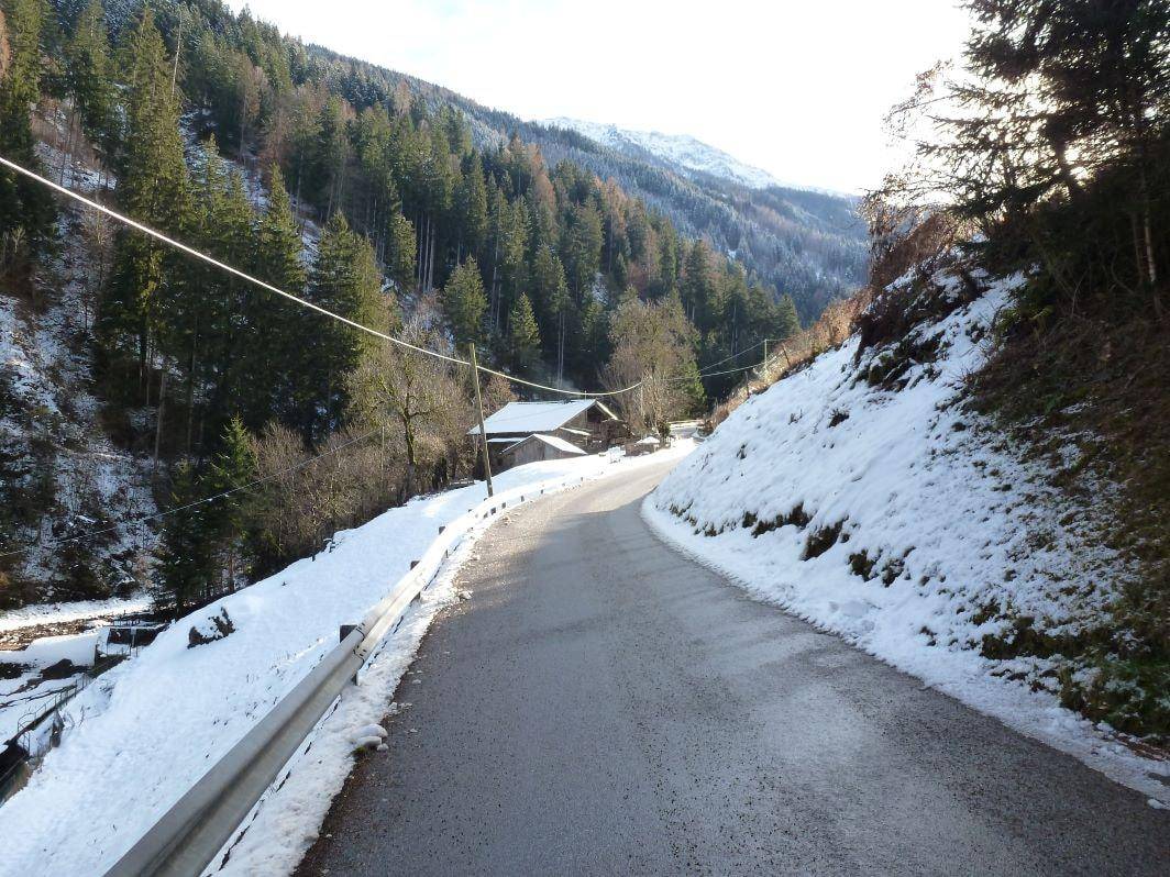 Verschneite Bergstraße führt durch eine winterliche Waldlandschaft zum abgelegenen Holzhaus in der Ferne.