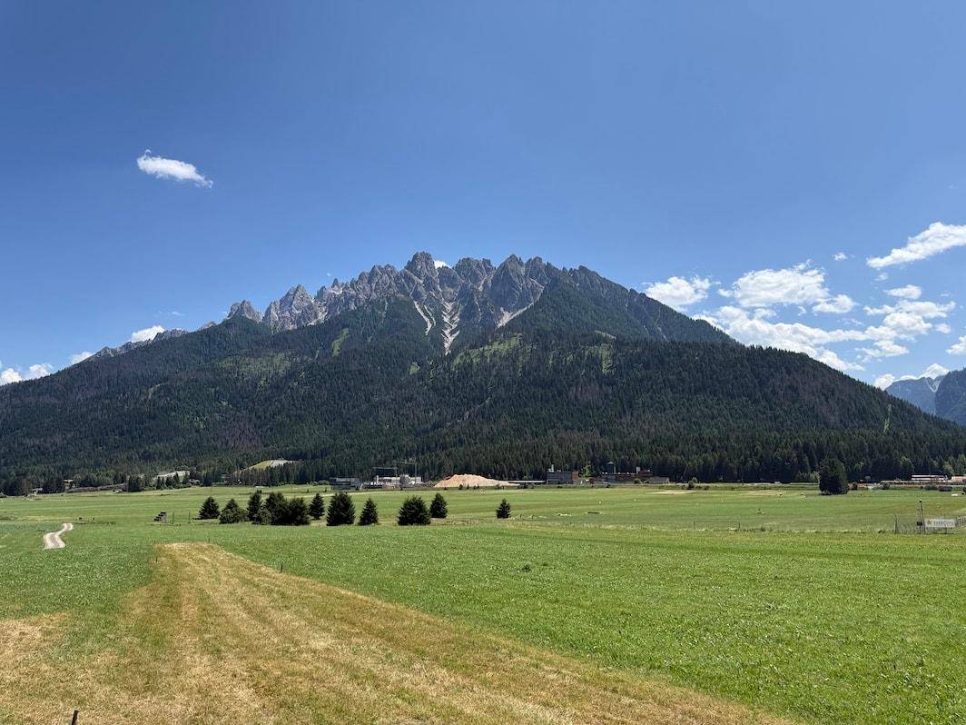Weite grüne Wiesen erstrecken sich vor einer imposanten Bergkette unter blauem Himmel.