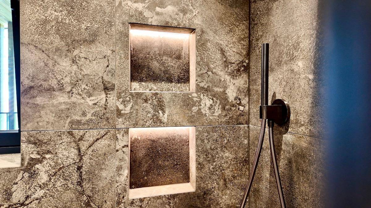 Detailansicht einer modernen begehbaren Dusche mit dunklen Fliesen und beleuchteten Nischen.
