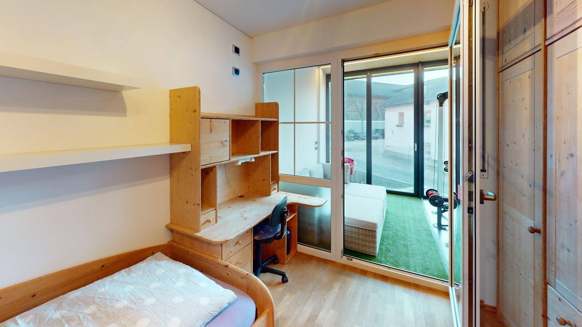 Helles Schlafzimmer mit Holzmöbeln, Schreibtisch und Zugang zu einem verglasten Balkon mit Kunstrasen.