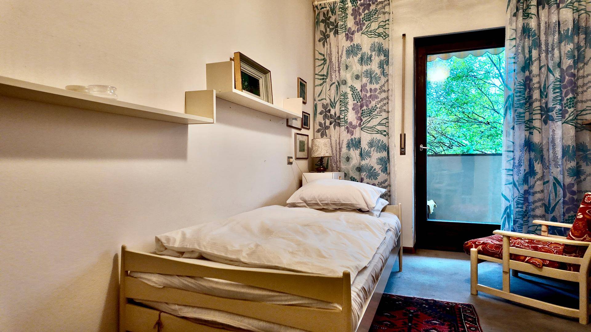 Helles Schlafzimmer mit Einzelbett, Teppichboden und Zugang zu einem kleinen Balkon.