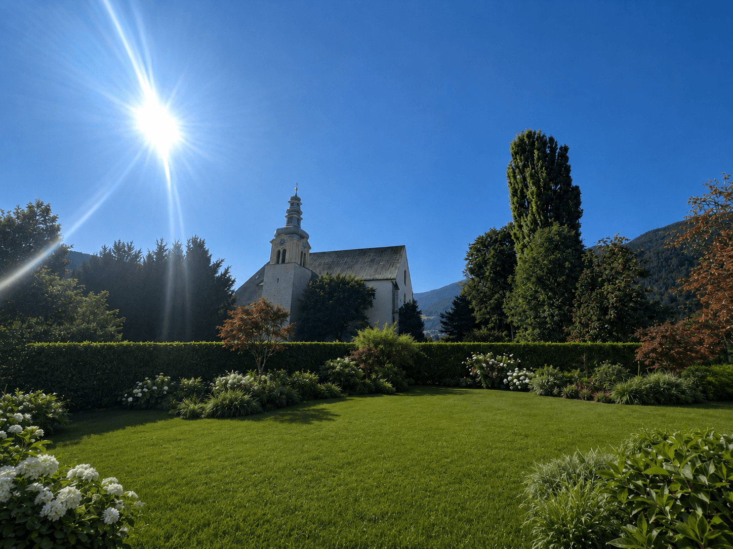 Gepflegter Garten mit Rasenfläche, Blumenbeeten und Blick auf eine historische Kirche im Hintergrund.