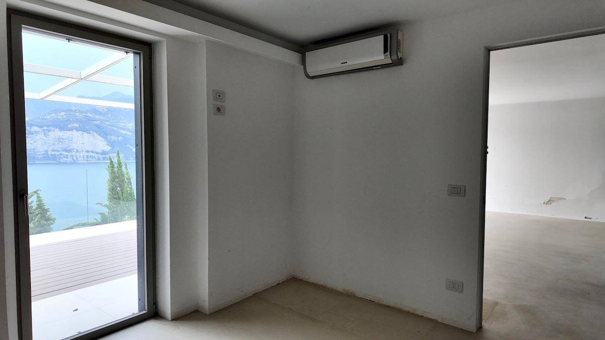 Leeres Zimmer mit Betonboden und Klimaanlage, mit Zugang zu einem Balkon und Blick auf den See.