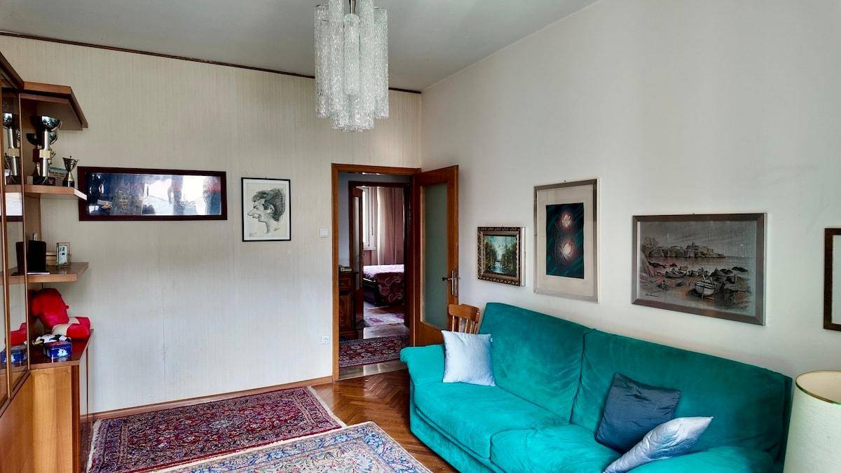 Geräumiges Wohnzimmer mit türkisfarbenem Sofa, Holzboden und Blick in einen weiteren Raum.