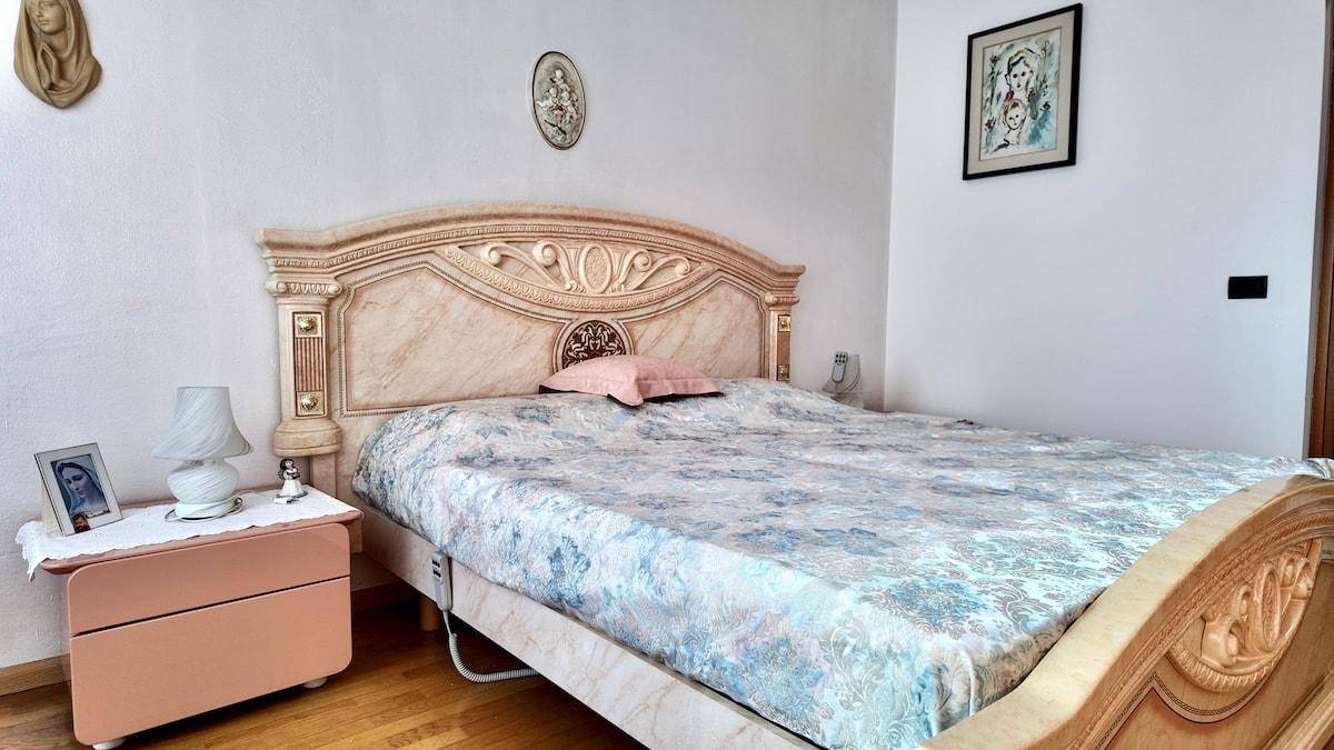 Schlafzimmer mit großem, reich verziertem Bett, Nachttisch und hellem Ambiente.