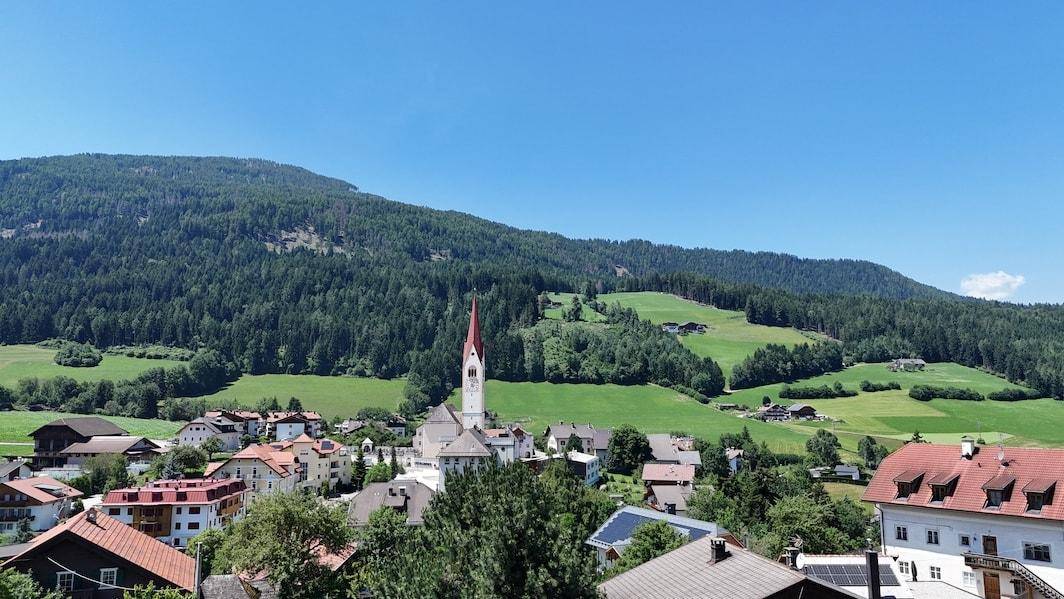 Weitläufiger Blick über ein idyllisches Dorf mit Kirchturm und bewaldeten Berghängen.