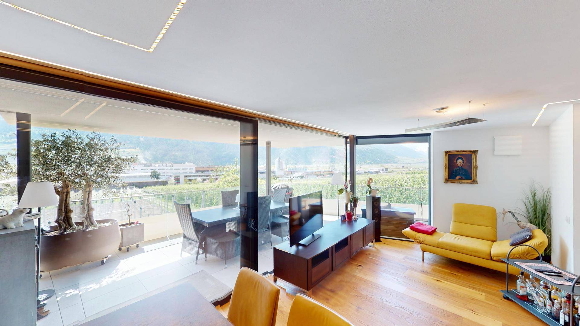 Gemütliches Wohnzimmer mit gelbem Sofa, Fernseher und Panoramablick auf Weinberge und Berge.