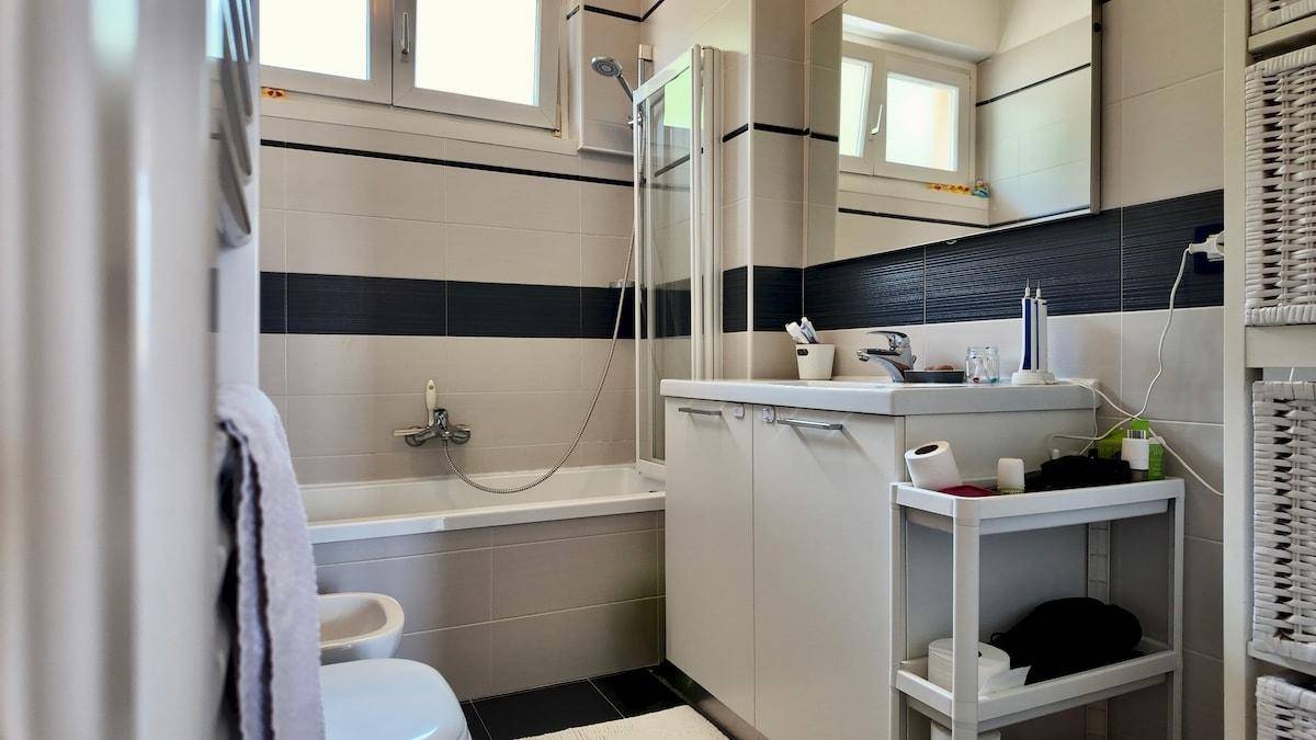Badezimmer mit Waschtisch, Spiegel, Badewanne und Duschvorrichtung, mit praktischen Ablagen.