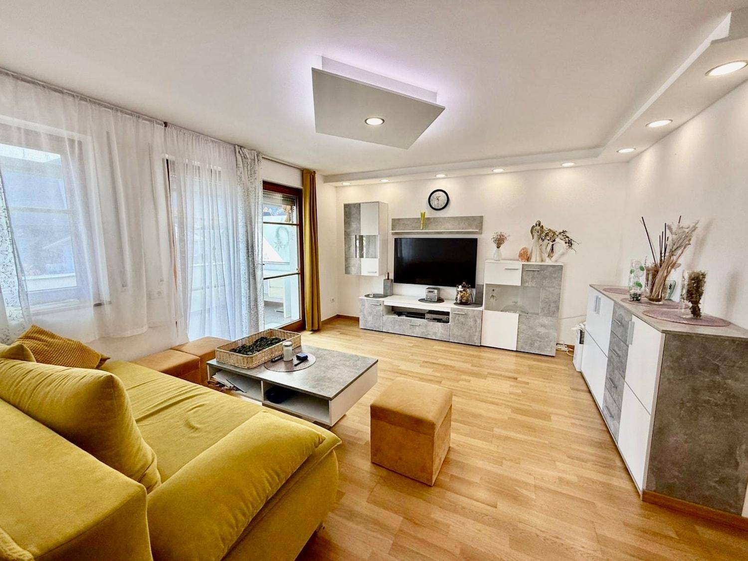 Geräumiges Wohnzimmer mit hellem Parkettboden, modernem TV-Möbel und Zugang zum Balkon.