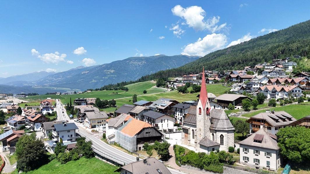 Luftaufnahme eines idyllischen Bergdorfes mit traditioneller Kirche, modernen Häusern und grüner Landschaft.