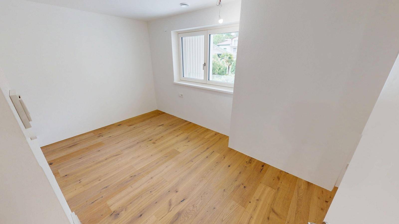 Helles Zimmer mit Parkettboden und einem Fenster, das viel Tageslicht hereinlässt.