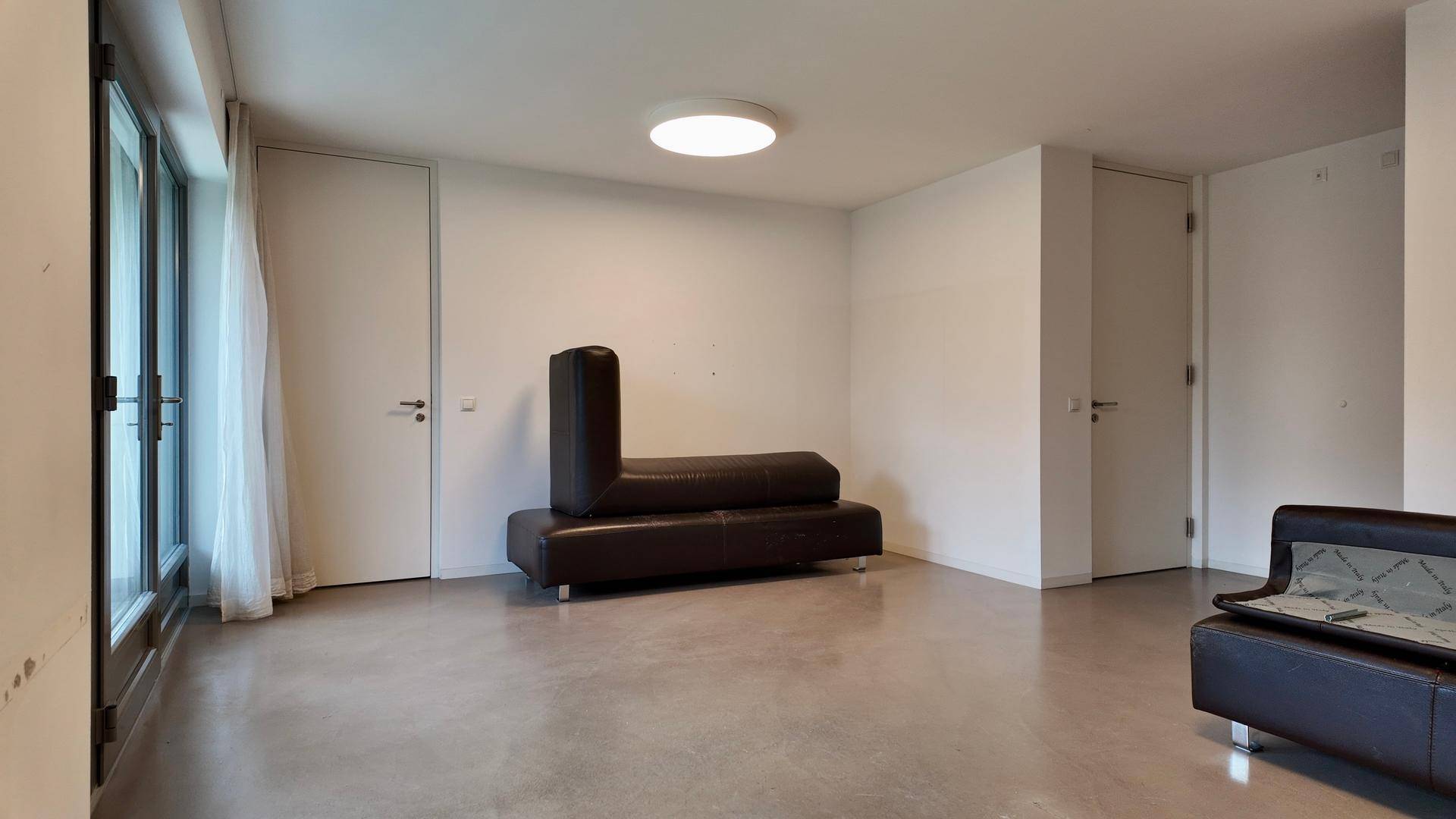 Heller Wohnraum mit einem dunklen Sofa, zwei weißen Türen und großen Fenstern, die viel Licht hereinlassen.