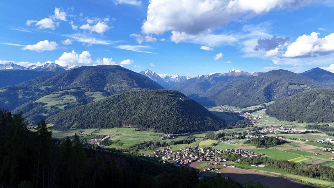 Weitläufige Berglandschaft mit grünen Tälern, bewaldeten Hügeln und kleinen Dörfern unter blauem Himmel.