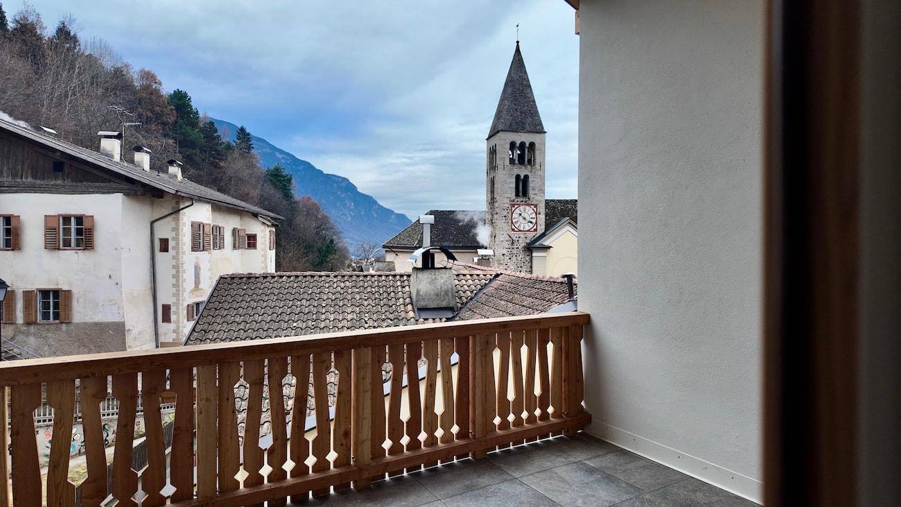 Balkon mit traditionellem Holzgeländer und malerischem Blick auf ein Dorf mit Kirchturm und Berge.