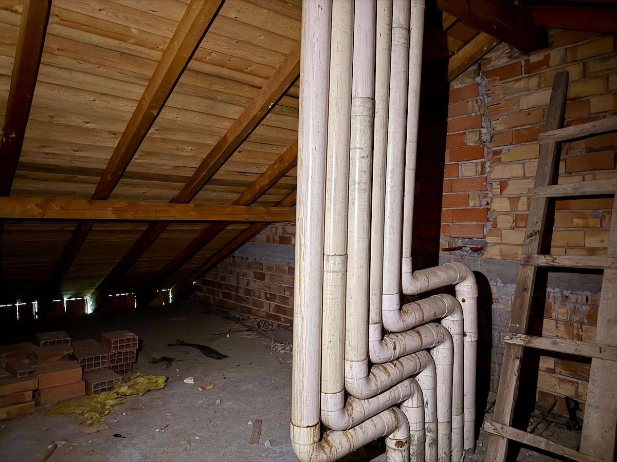 Dachboden mit sichtbaren Holzbalken, Ziegelwänden und vertikalen Rohren, bietet Stauraum.