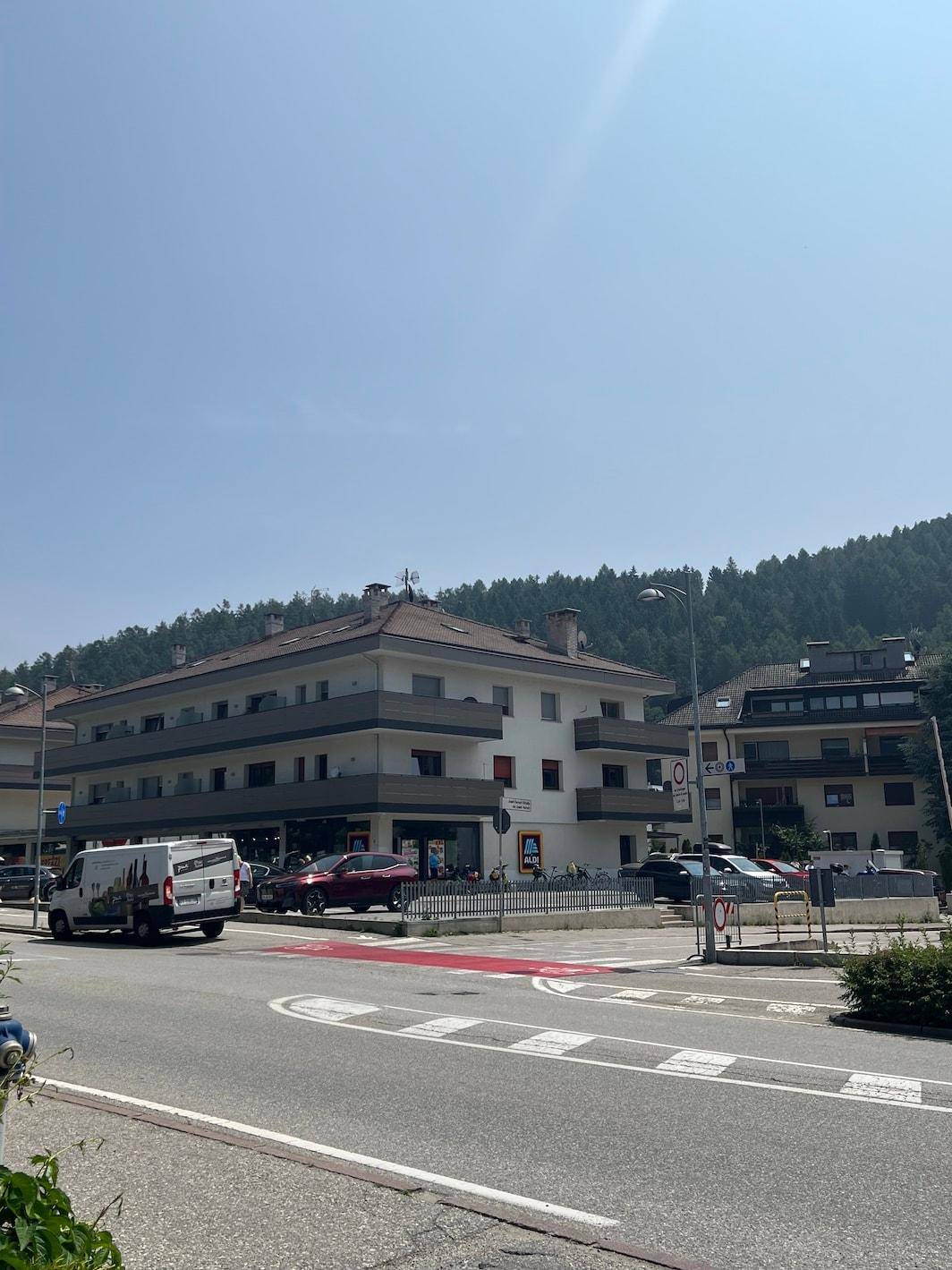 Gebäudeansicht mit Supermarkt und Wohnungen, umgeben von grünen Hügeln unter blauem Himmel.