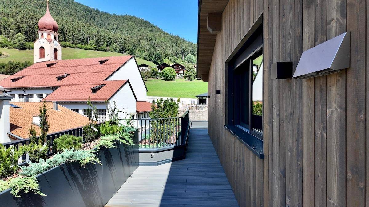 Terrasse mit Holzboden und Pflanzen, bietet einen charmanten Blick auf das Dorf mit Kirchturm und grüne Hügel.