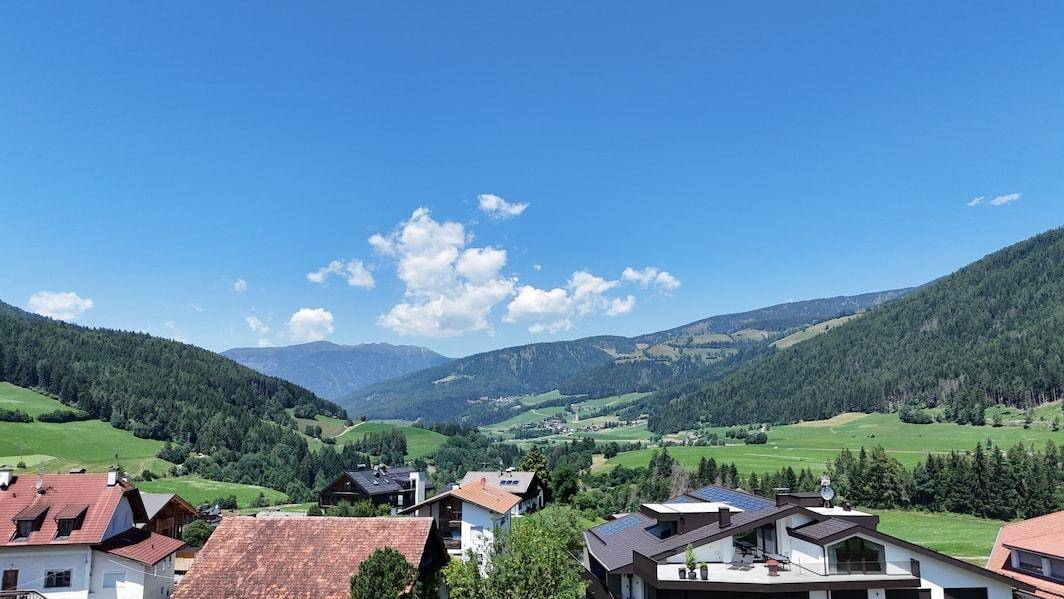 Panoramablick über ein grünes Tal mit Häusern, Feldern und majestätischen Bergen unter blauem Himmel.
