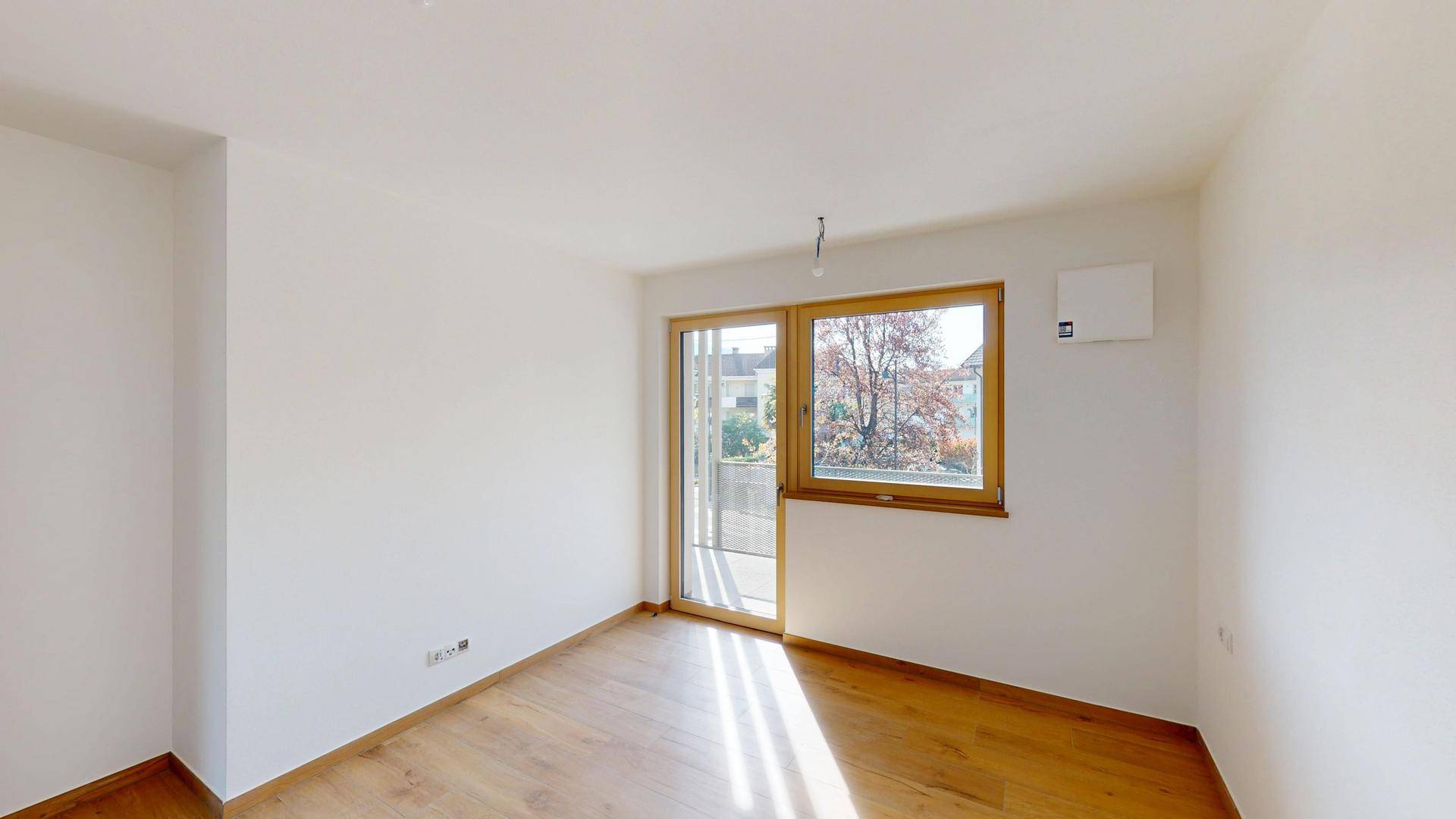 Unmöbliertes Zimmer mit hellem Holzboden und Fenster, das viel Tageslicht hereinlässt.