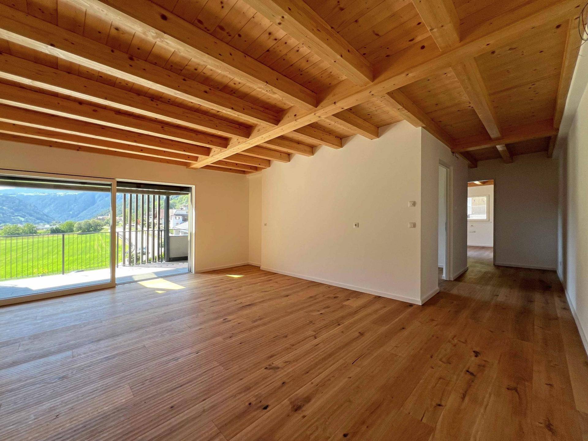 Heller Wohnraum mit Holzboden, sichtbaren Deckenbalken und Zugang zu einem Balkon mit Bergblick.