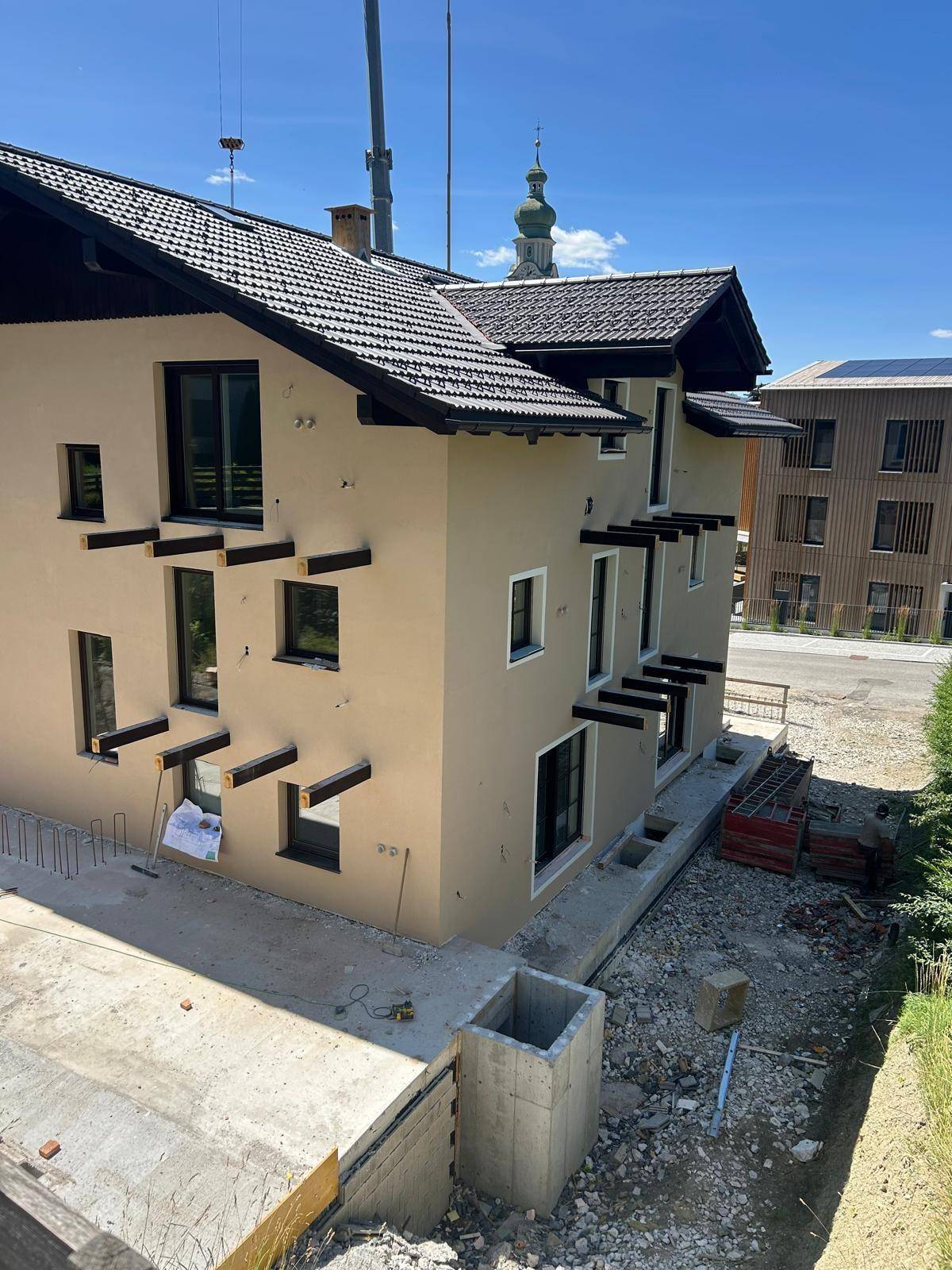 Neubauhaus in alpiner Umgebung mit beiger Fassade und dunklem Dach, umgeben von Baustelle.