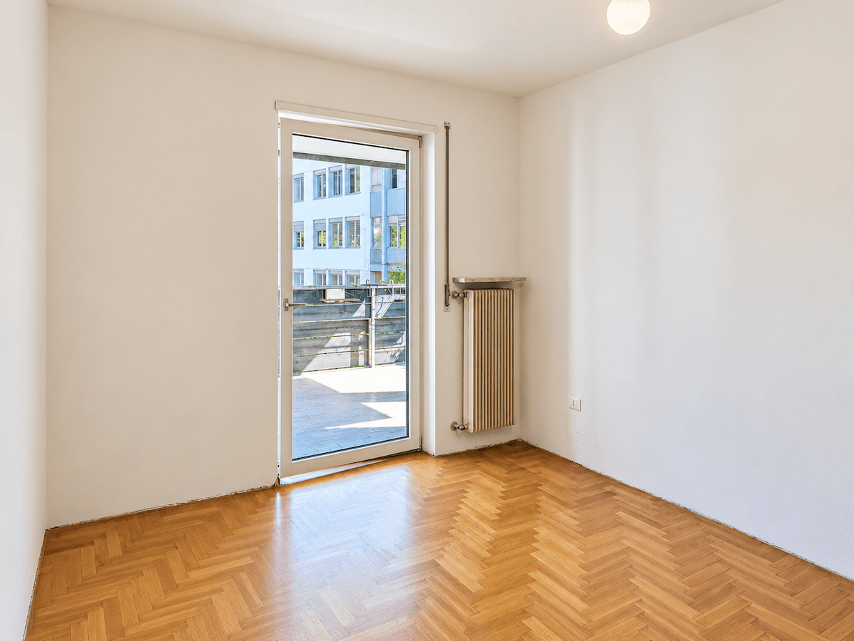Helles Zimmer mit Fischgrätparkett, Heizkörper und Zugang zu einem kleinen Balkon.