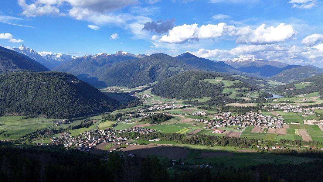 Weitläufige Berglandschaft mit grünen Tälern, bewaldeten Hügeln und kleinen Dörfern unter blauem Himmel.