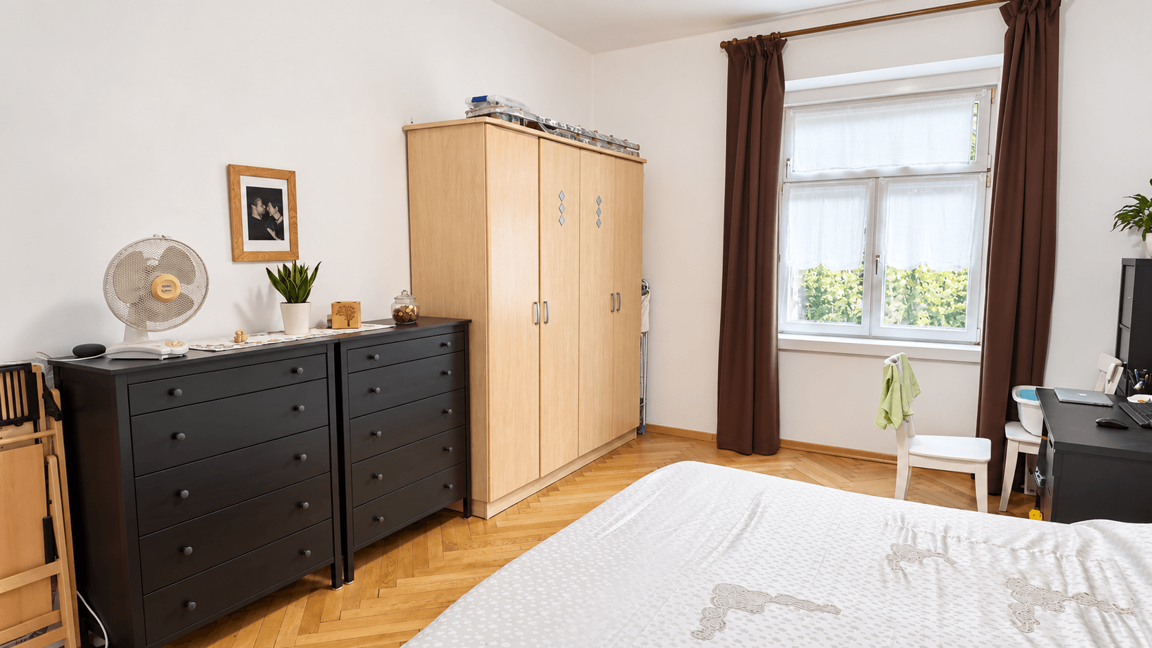 Schlafzimmer mit Holzboden, Kommoden und einem Fenster mit Blick ins Grüne.