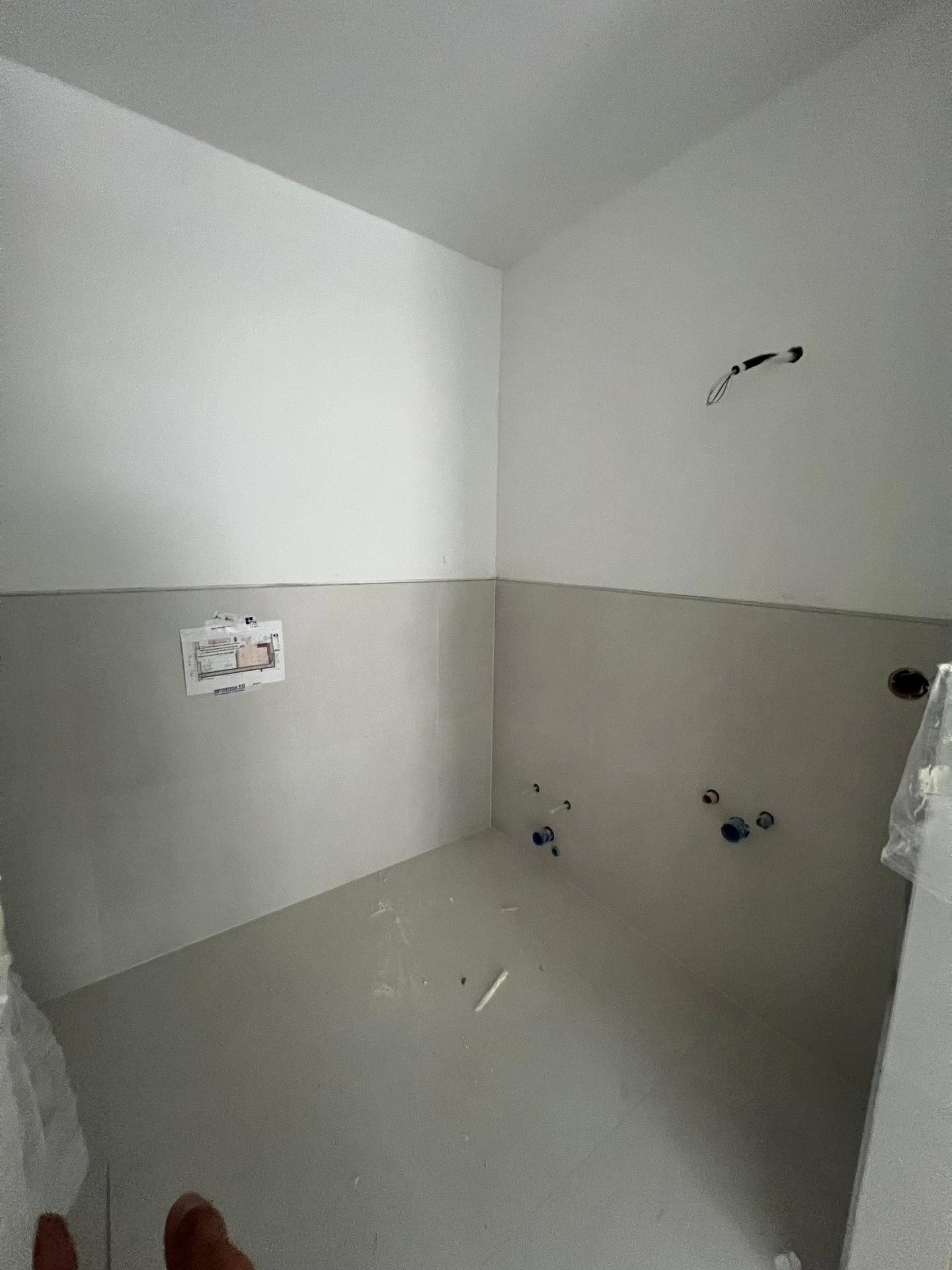Rohbau-Badezimmer mit vorbereiteten Anschlüssen, hellen Wänden und Betonboden.