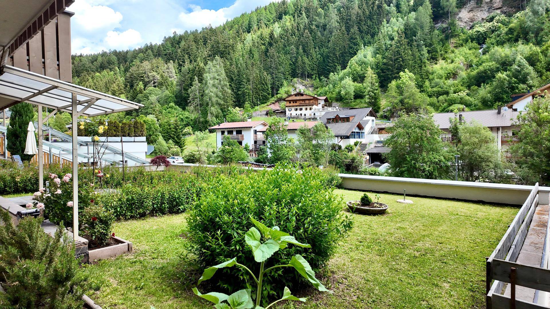 Weitläufiger Garten mit Rasen und Sträuchern, umgeben von Häusern und einer beeindruckenden Berglandschaft.