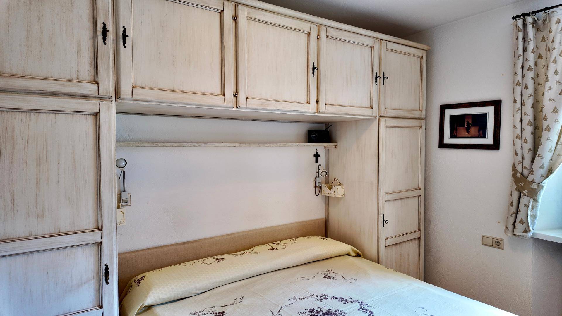 Gemütliches Schlafzimmer mit integrierten, hellen Holzschränken über dem Bett und einem Fenster mit gemusterten Vorhängen.