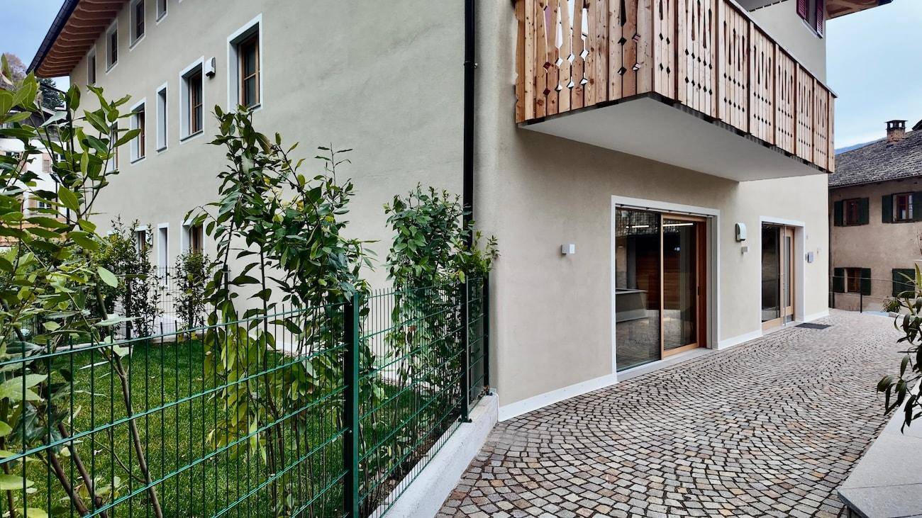 Seitenansicht eines Gebäudes mit Holzbalkon und gepflastertem Vorplatz, umgeben von Grün.