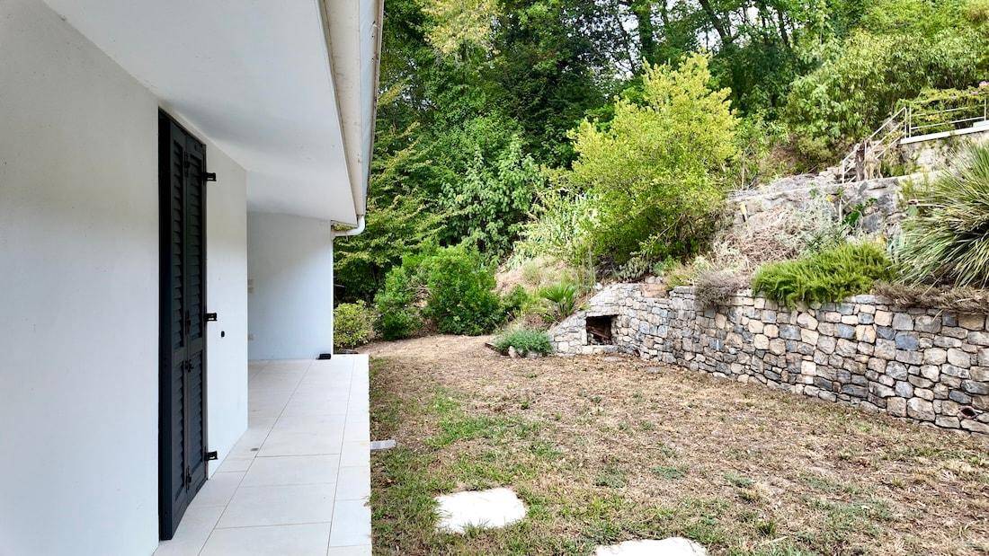 Gartenbereich neben einem weißen Haus mit Trockenrasen, Steinmauer und üppigen Bäumen am Hang.