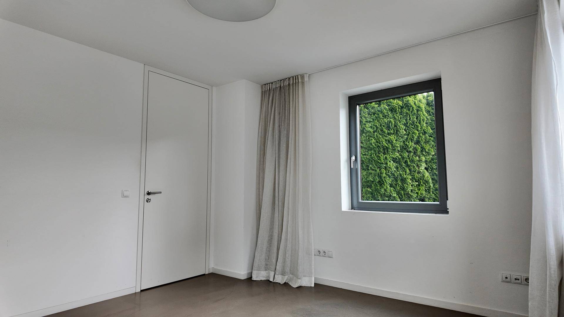 Helles Schlafzimmer mit einem Fenster, das einen Blick ins Grüne bietet, und einer schlichten weißen Tür.