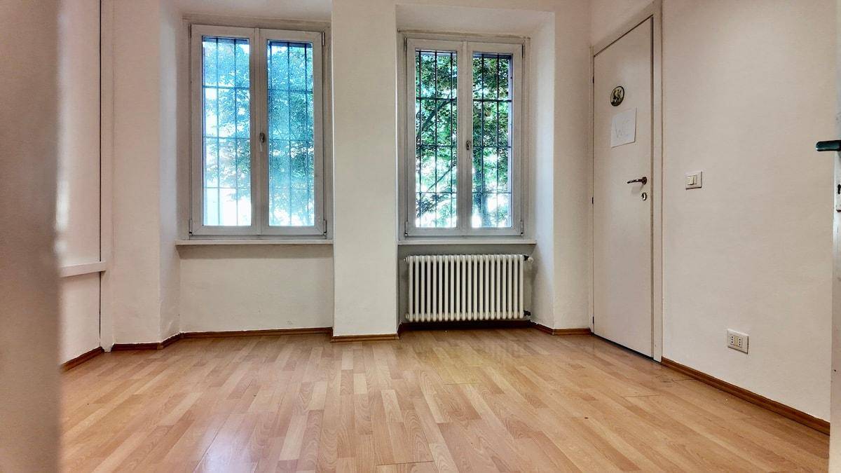 Leerer Raum mit Holzboden, weißen Wänden und zwei Fenstern mit Blick auf Bäume.