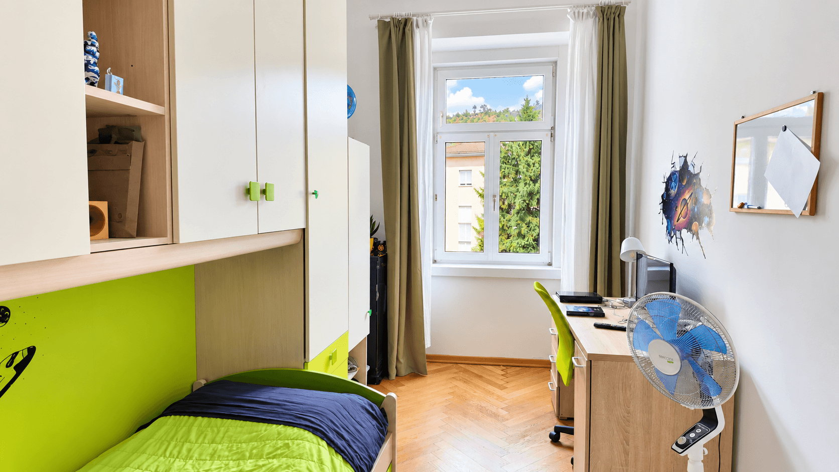 Kinderzimmer mit Schreibtisch, Einzelbett und Blick ins Grüne durch das Fenster.