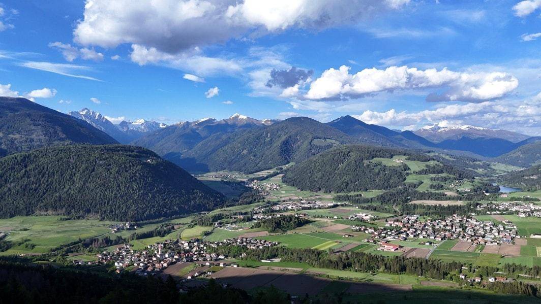 Weitläufige Berglandschaft mit grünen Tälern, bewaldeten Hügeln und kleinen Dörfern unter blauem Himmel.